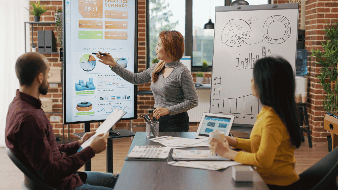 Businesswoman presenting analytics data on a large screen while two team members review charts and graphs during a meeting in a modern office setting.