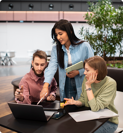 Three young professionals collaborating at a laptop in a modern open workspace, reviewing notes and discussing ideas.