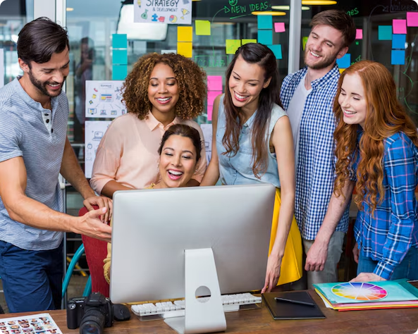Group of young professionals gathered around a desktop computer, smiling and collaborating in a colorful, creative office space with sticky notes on the glass wall.